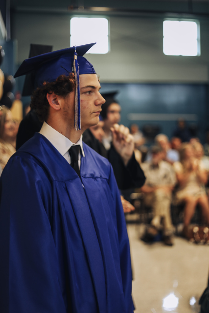 A young man in a blue gown and cap stands in a room; an audience sits behind him.