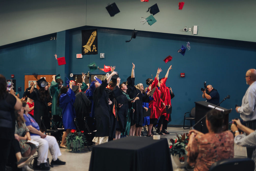 A group of people in gowns and hats throw them in the air at an indoor event.