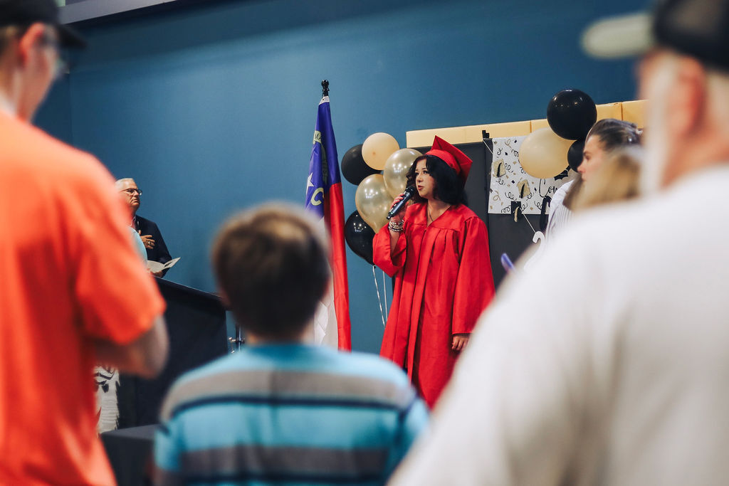 A person in a red graduation gown speaks into a microphone at a graduation ceremony.