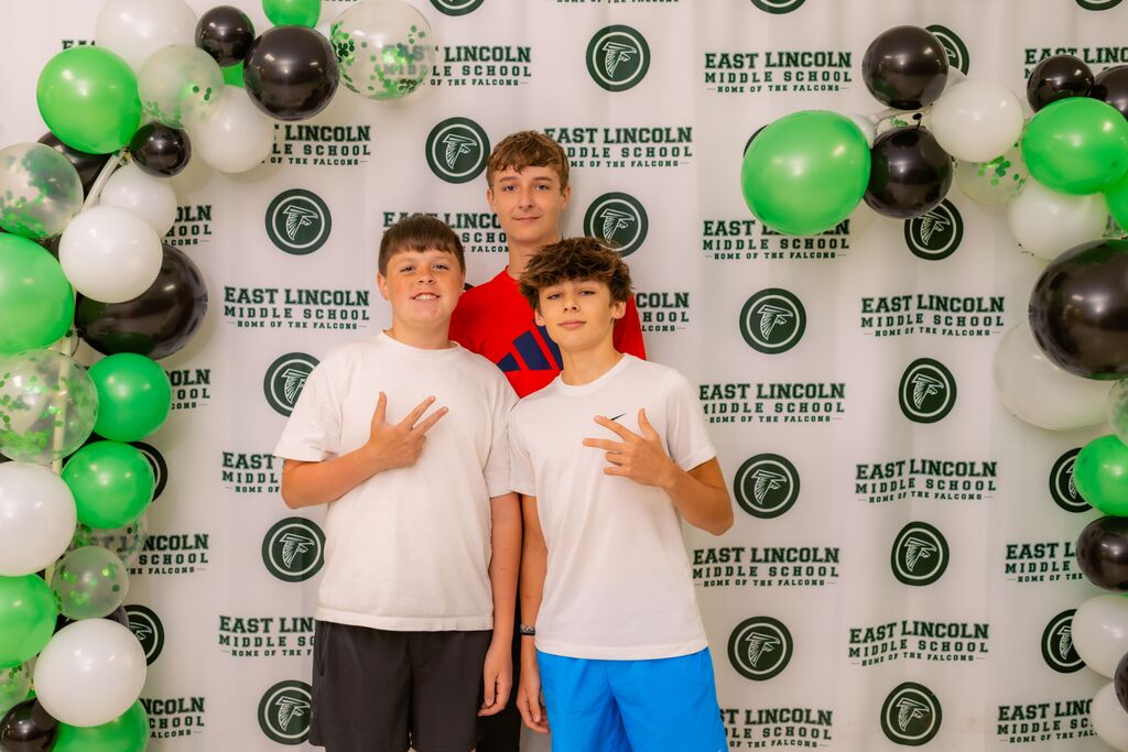 Three boys in white t-shirts, posing for a photo in front of a backdrop with green and black balloons.