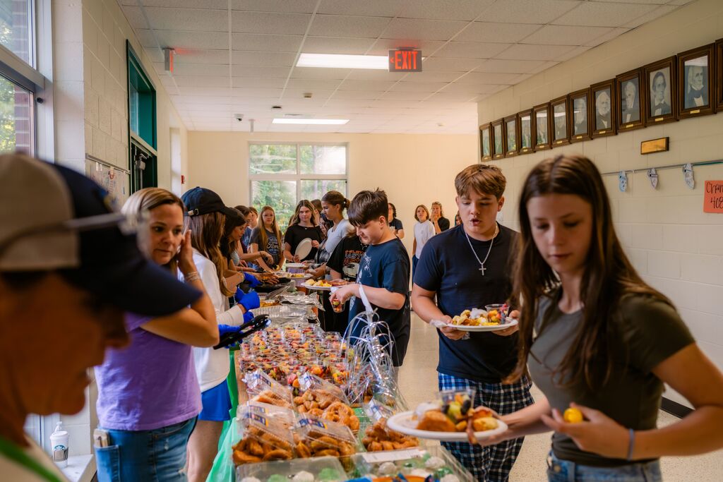 A long buffet table with food, many people wearing casual clothing, lined up and holding plates.