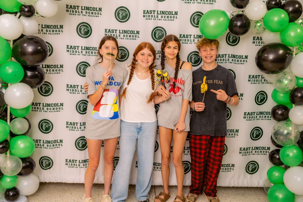 Four people stand in front of a wall with balloons and a banner, smiling and posing for a photo.