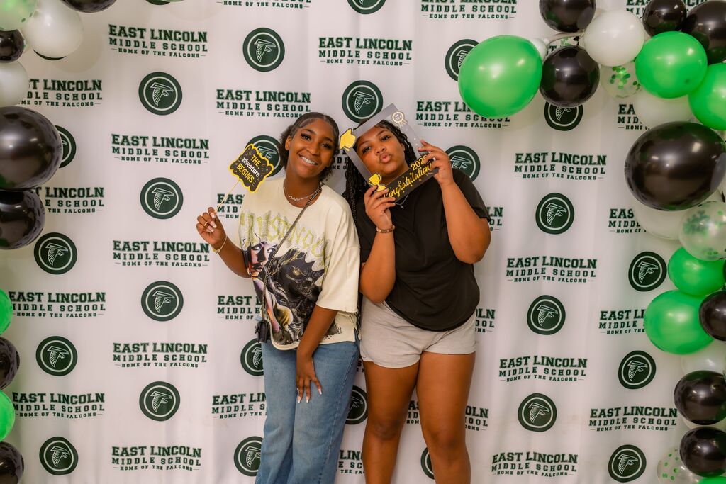 Two individuals stand in front of a green-and-black balloon backdrop with white text. Both hold up objects and smile.