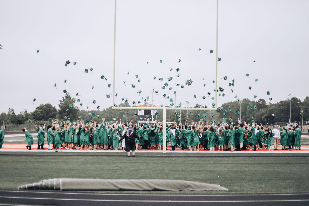 Graduating students in green gowns throw caps into the air on a football field with trees in the distance.