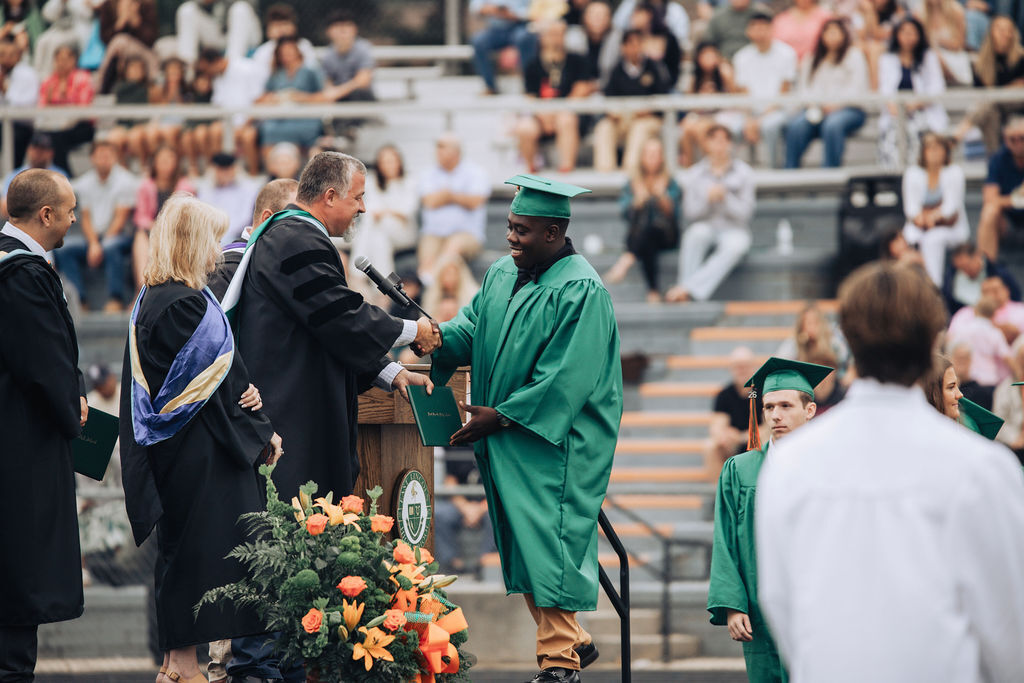 A man in a green graduation gown shakes hands with a faculty member on stage. A crowd sits on bleachers.