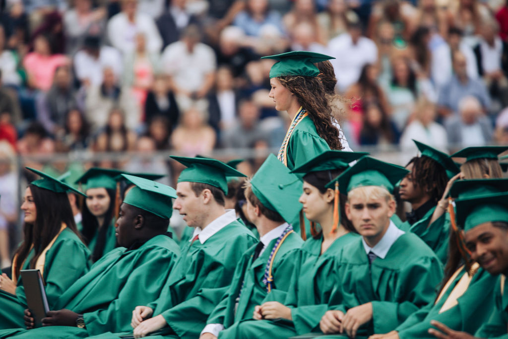 Graduates wearing green gowns and caps sit in rows with audience in stadium.