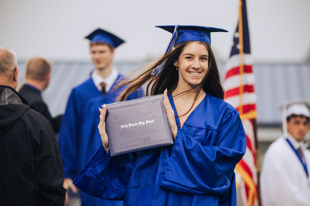 Woman in blue graduation attire holds her diploma and smiles at the camera. Other people in caps and gowns stand behind her, and an American flag is in the background.