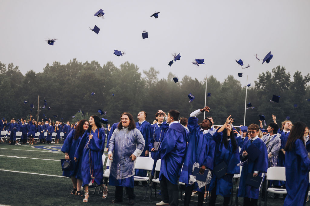 Graduating students on a field, throwing their caps in the air. Trees and a cloudy sky in the background.
