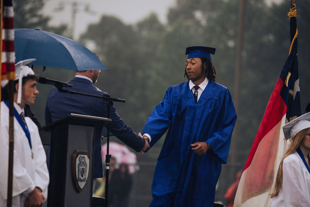 A person in a blue graduation gown shakes hands with a speaker at a podium. Two flags stand behind them.