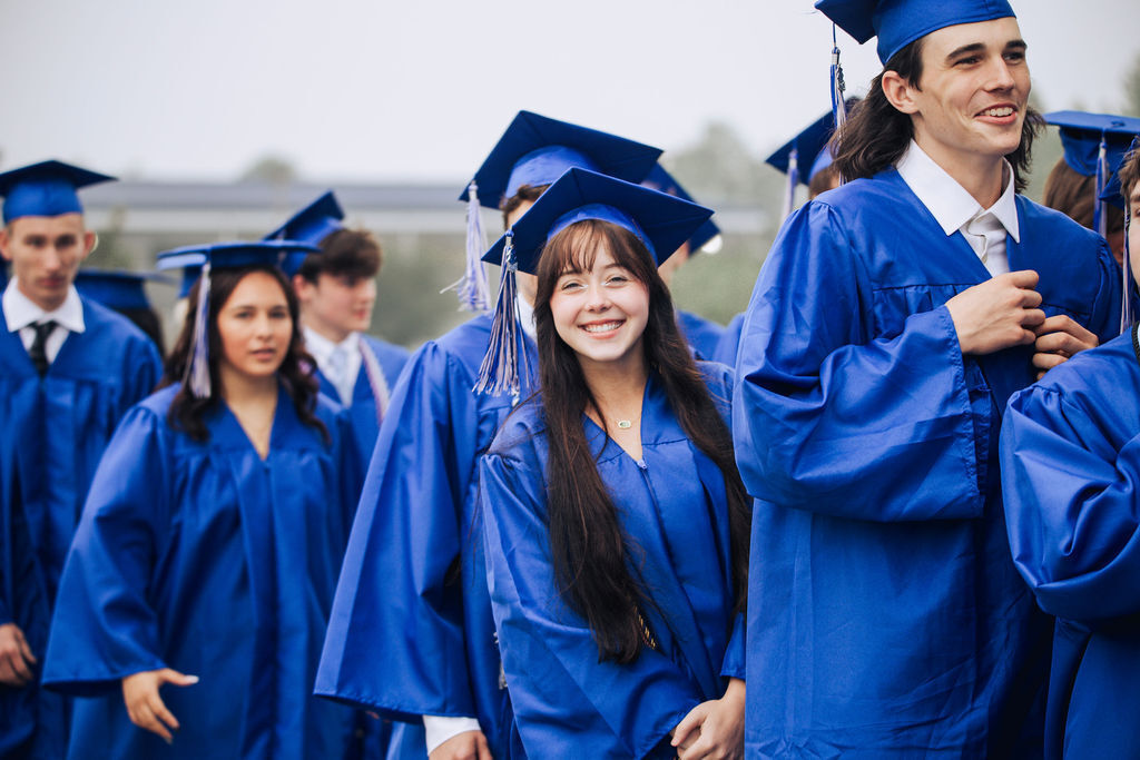 Graduates in blue caps and gowns stand together, smiling. A woman with long hair smiles and looks at the camera.