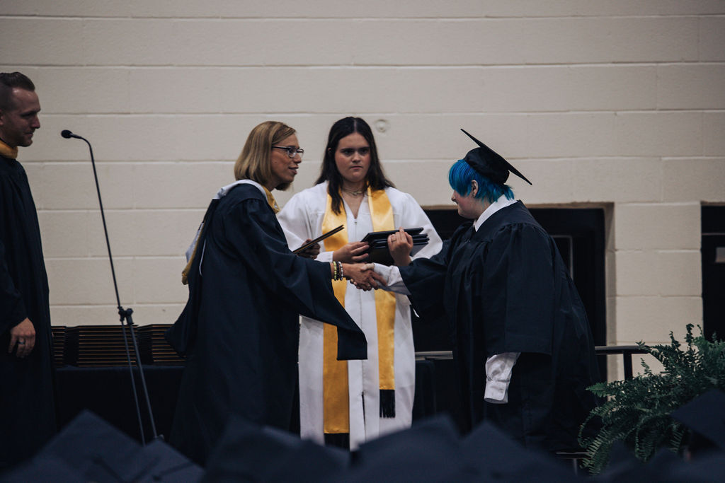A group of people in academic robes and hats stand on a stage. A person receives an item from another.