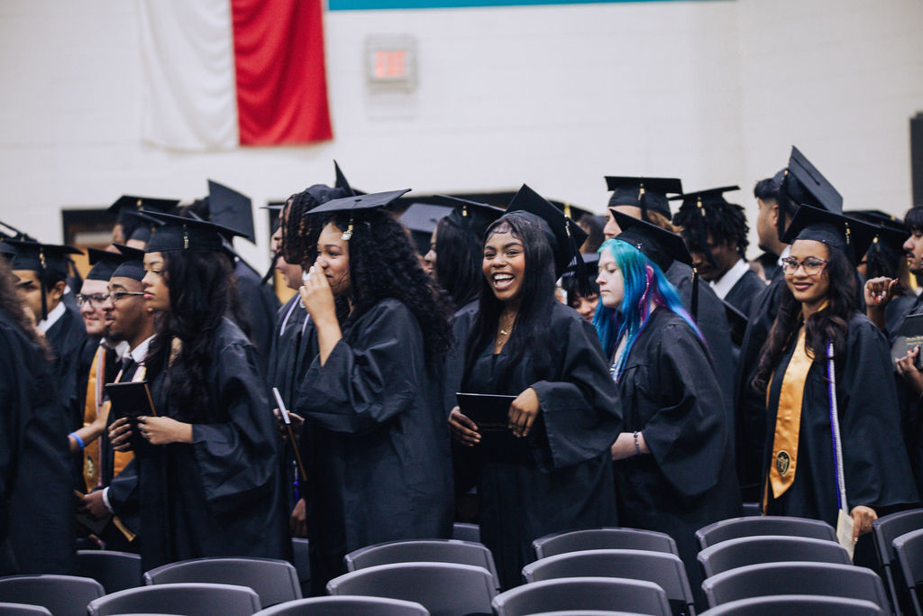 Graduates in black gowns and caps stand in rows, some smiling, at a graduation ceremony. A flag hangs on the wall.