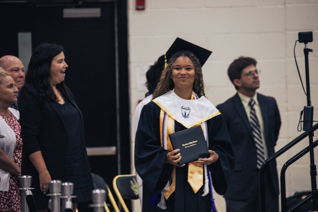 A woman in a cap and gown holds a diploma. Behind her, a man in a suit stands near a staircase.