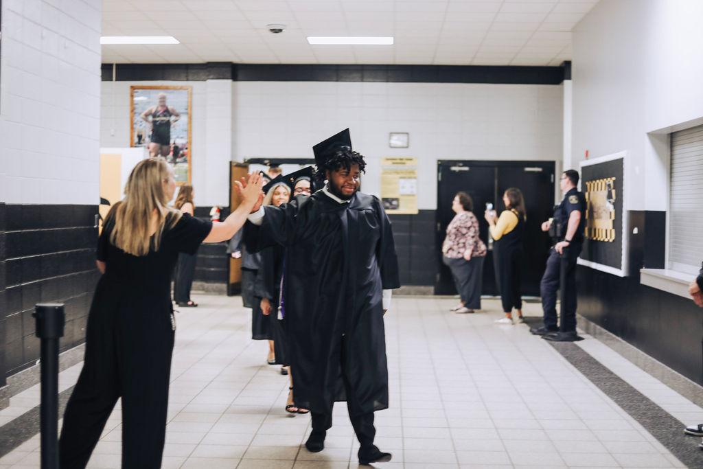 People gather in a hallway with white walls and a black border. A man in a cap and gown holds his cap.