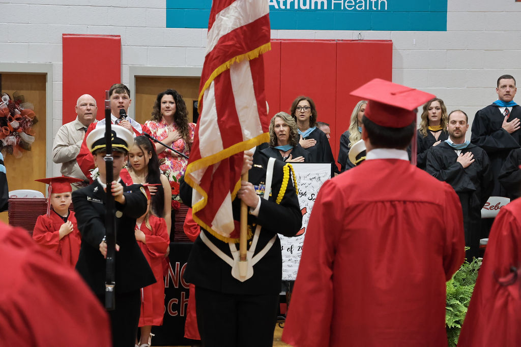 Several people in red gowns stand in a room with a flag, some wearing caps. A banner in the background reads Atrium Health.