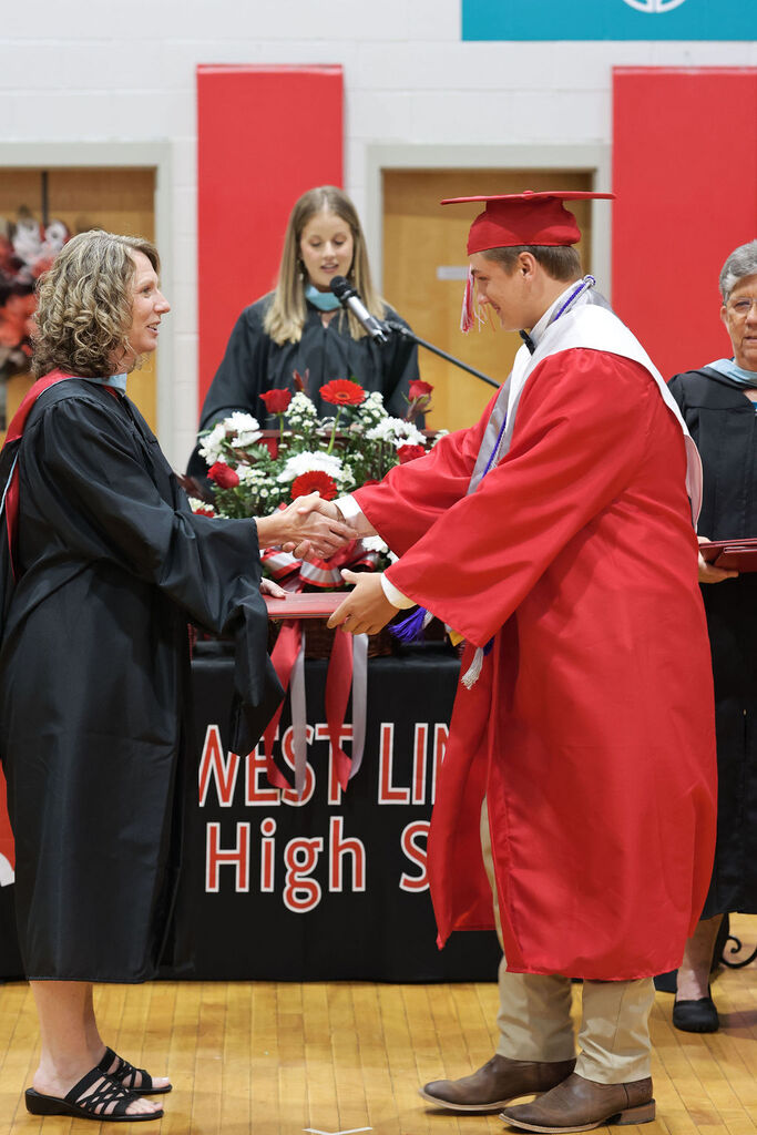 A man in a red cap and gown shakes hands with a woman in a black gown. They stand in front of a table with flowers and a banner reading "West Lin High S".