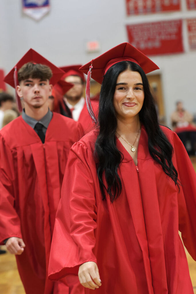 Graduates in red gowns and caps, smiling, walking in a school gym. Red banners adorn the background.