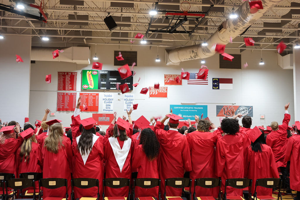 Graduates in red gowns and caps throwing caps into the air during a ceremony in a gym.