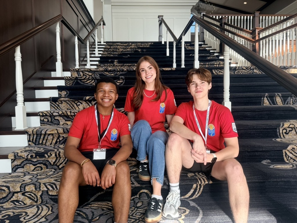 Three individuals in red shirts sit on carpeted stairs. The stairs have a geometric pattern, white railings, and a white wall.