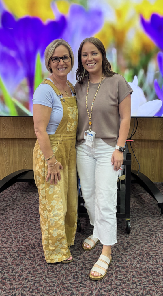 Two women stand smiling in front of a large digital display showing vibrant purple and yellow flowers. The woman on the left wears yellow floral overalls over a light blue shirt, and the woman on the right wears a tan short-slee shirt and white cropped pants. 