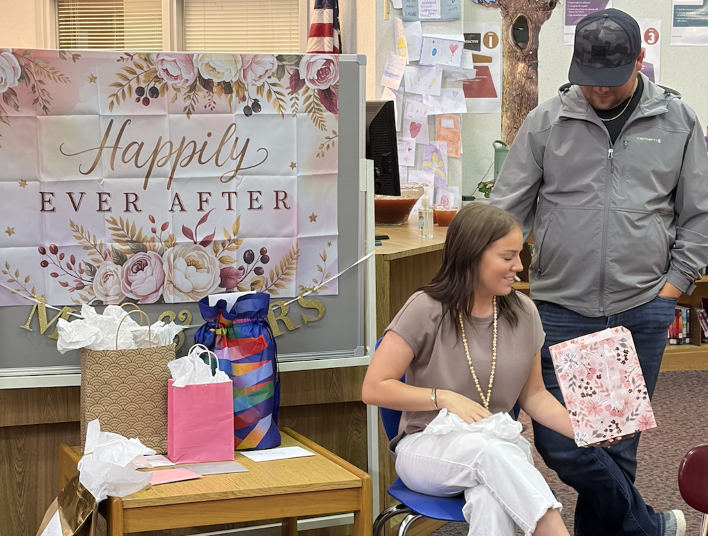 A woman in a tan shirt and white pants sits in a blue chair, smiling as she holds a pink floral gift bag. A man in a grey Carhartt jacket and camouflage hat stands behind her. To their left is a table with several gift bags and a large sign with floral borders that reads "Happily EVER AFTER" in gold lettering. The setting is a school media center with student work visible in the background.
