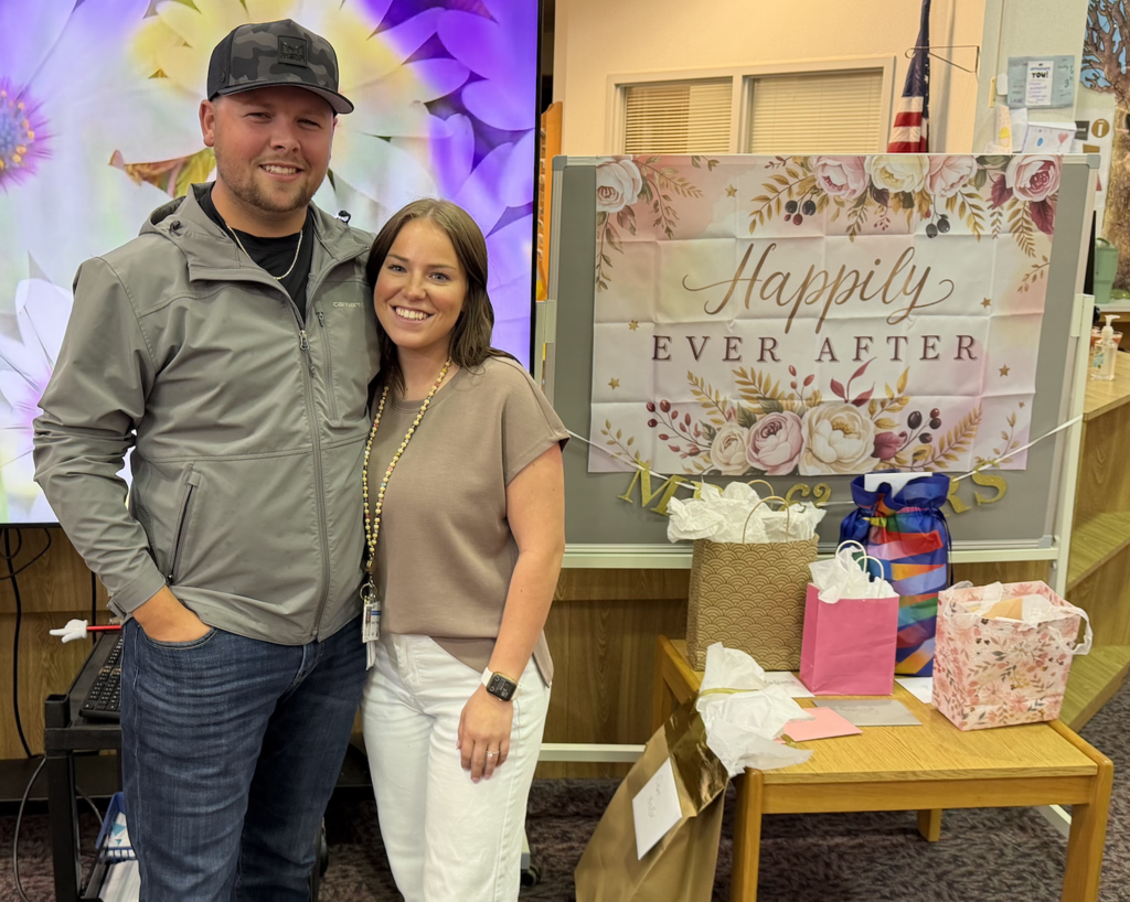 A man in a grey jacket and camouflage hat stands next to a woman in a tan shirt and white pants in a school media center. Behind them is a large screen displaying purple flowers and a sign that reads "Happily Ever After." A small wooden table to the right holds several gift bags and cards.