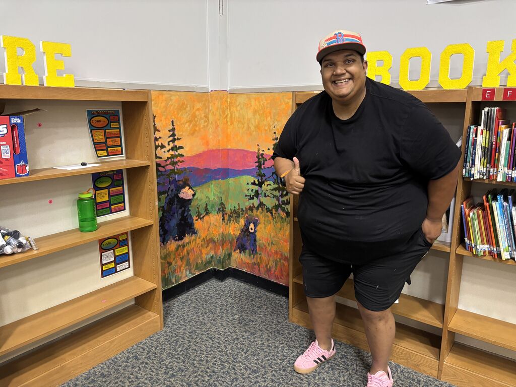 A smiling man giving a thumbs-up stands in a school library corner next to a painted wall mural. The colorful mural depicts two black bears in a grassy mountain landscape under an orange sunset. Wooden bookshelves flank the corner, with large yellow letters spelling parts of words on top of them.