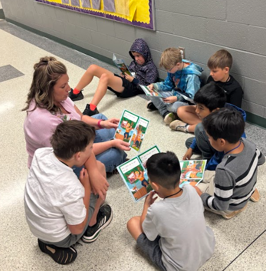 A woman sits with a group of young boys on the floor, reading books. Yellow wall decoration behind them.