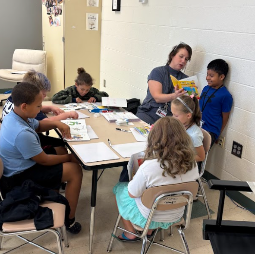 A teacher is reading a book to a group of children sitting at a table in a classroom.