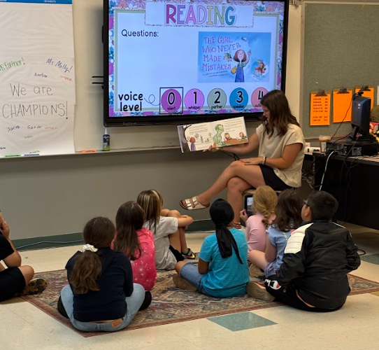 A woman reads a book to a group of children sitting in a circle on a rug.
