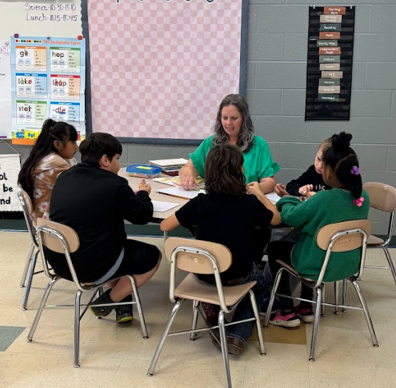 A group of children seated around a table with a teacher. The table has papers and a book.