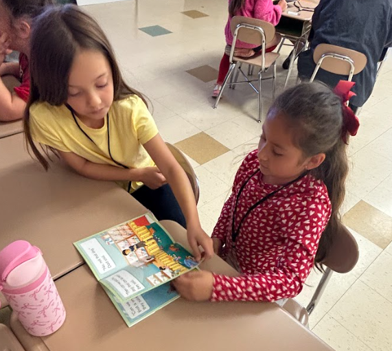Two young girls, one reading a book aloud to the other, seated at a table in a classroom.