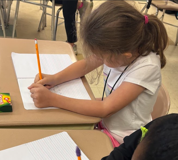 A child sits at a desk holding a pencil, working on a notebook. Another child is seated beside them, looking down.