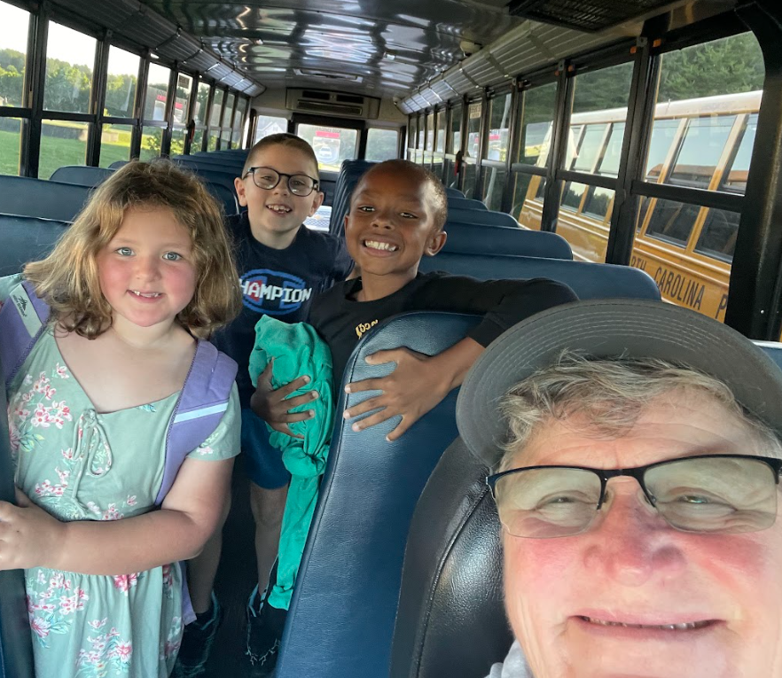 A school bus interior with three children and an adult. The adult has a hat and glasses.