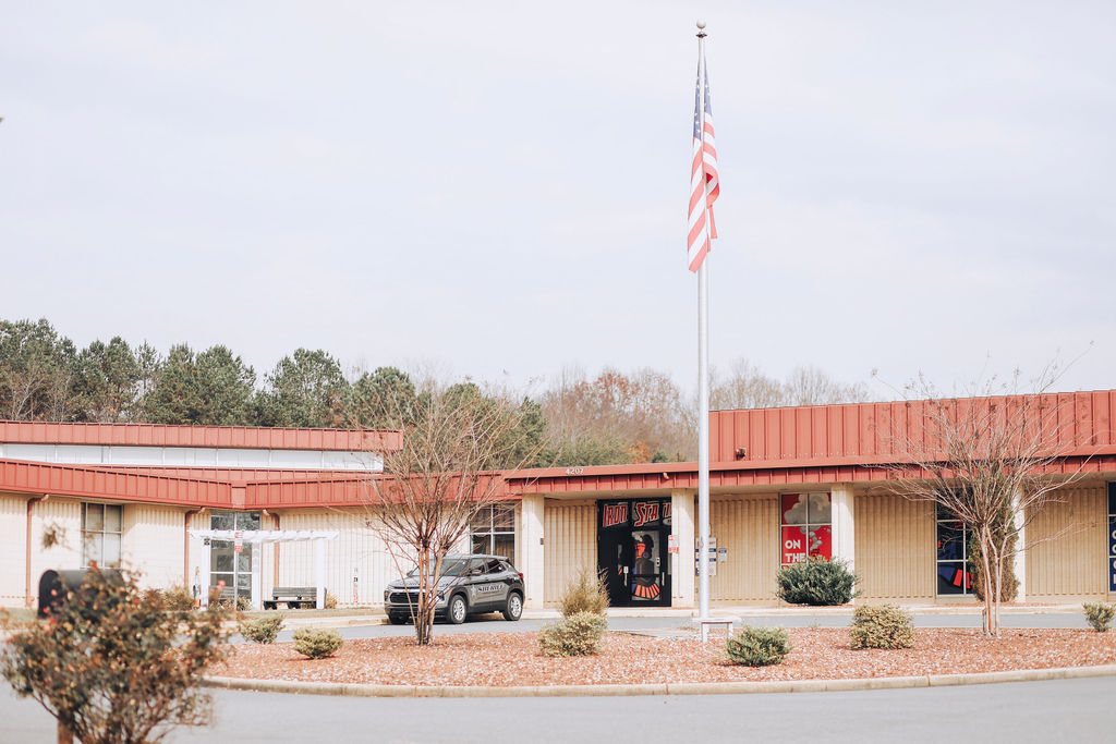A building with red roofing and a flagpole in front. A car is parked outside, with trees and plants around it.