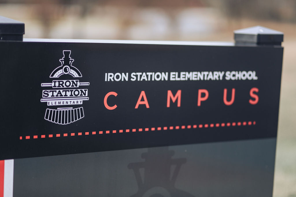 A black sign with a white logo and the words "Iron Station Elementary School Campus" in red. Background is blurred.