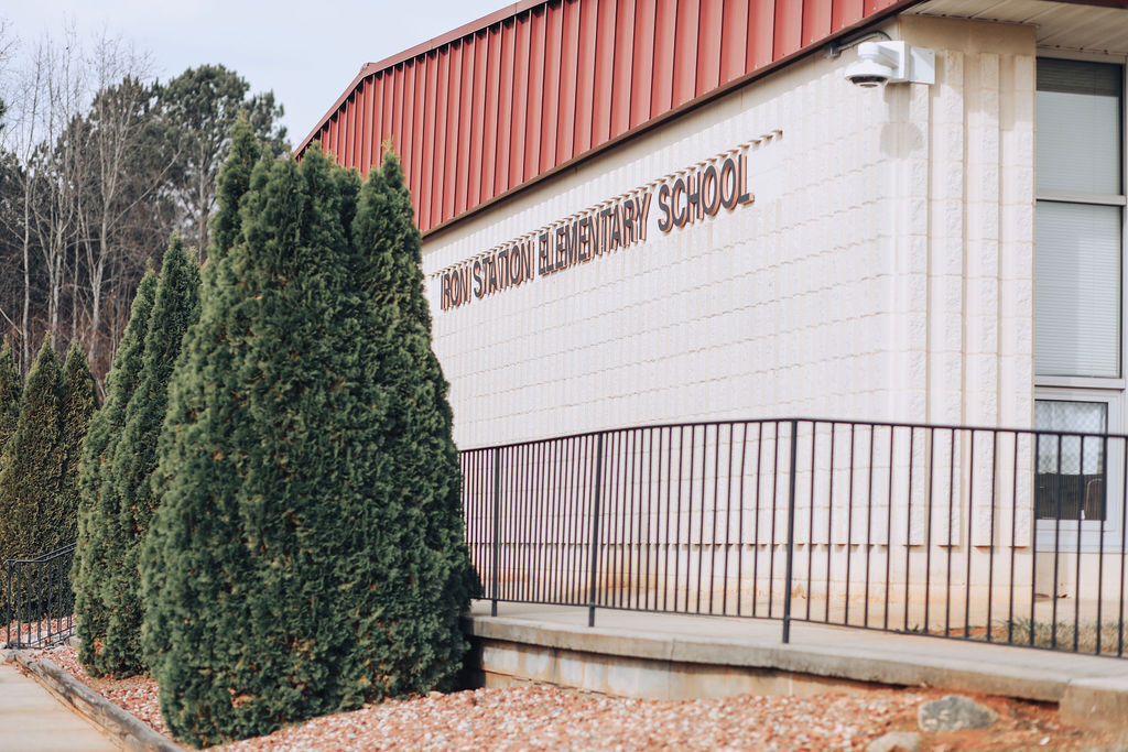 A school building with a red roof and white walls, "High School Elementary School" sign. Black railing on sidewalk. Green trees in front.