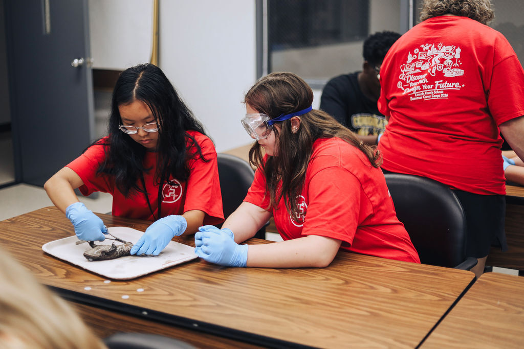 Two students in lab coats and gloves work on a fossil at a table in a classroom.