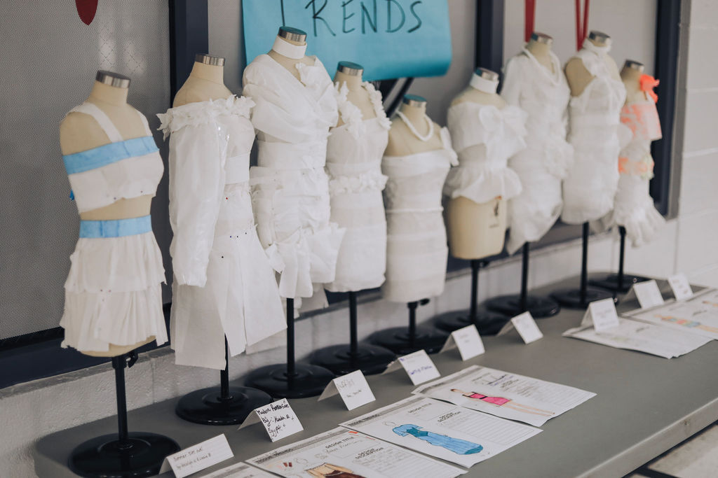 Mannequins displaying white dresses on stands with blue and white tags. An open book and papers on a table.