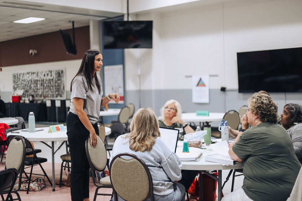 A woman stands before a group of seated individuals in a room with white walls, tables, and a television.