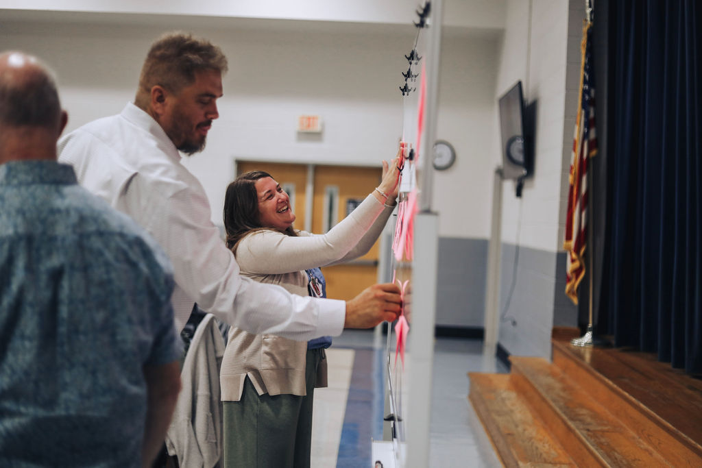 Three people standing in a room. A woman pins a pink paper to a bulletin board.