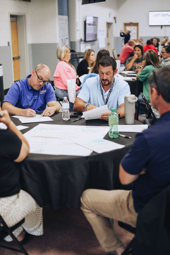 A group of people sit around a table, with papers and pens. A person holds a bottle of water.