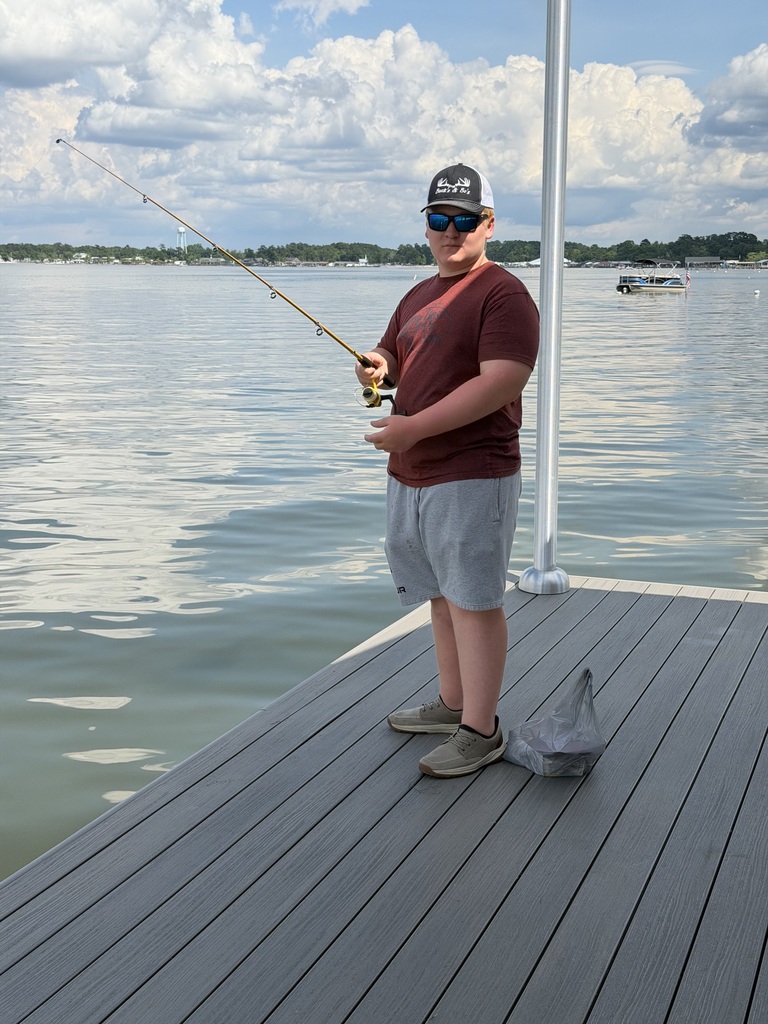 A person stands on a wooden dock by the water, holding a fishing rod and wearing sunglasses.