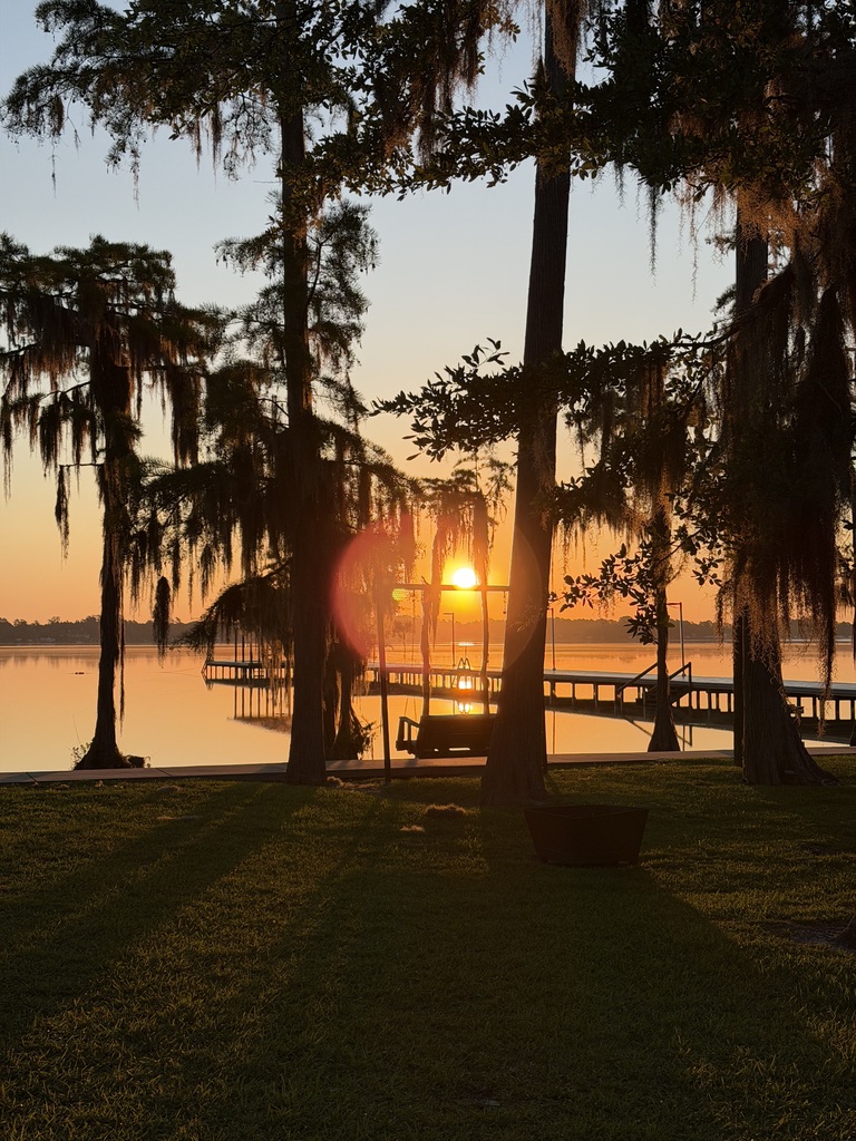 Sunset over water with dock, surrounded by trees and grass, creating a serene landscape.