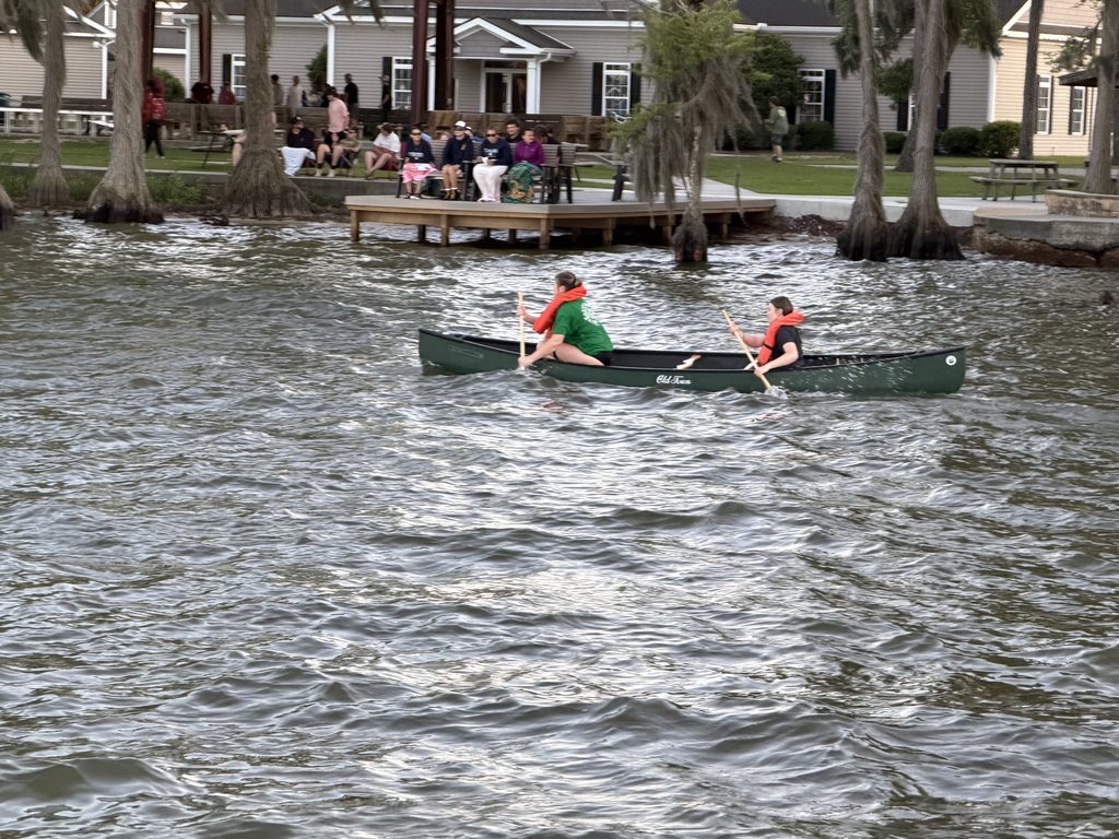 Two people in a green canoe paddle across the water with a pier and homes in the background.