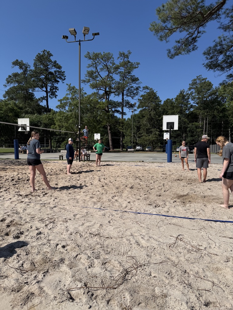 People play volleyball in a sandy area with trees. Two volleyball nets are set up.