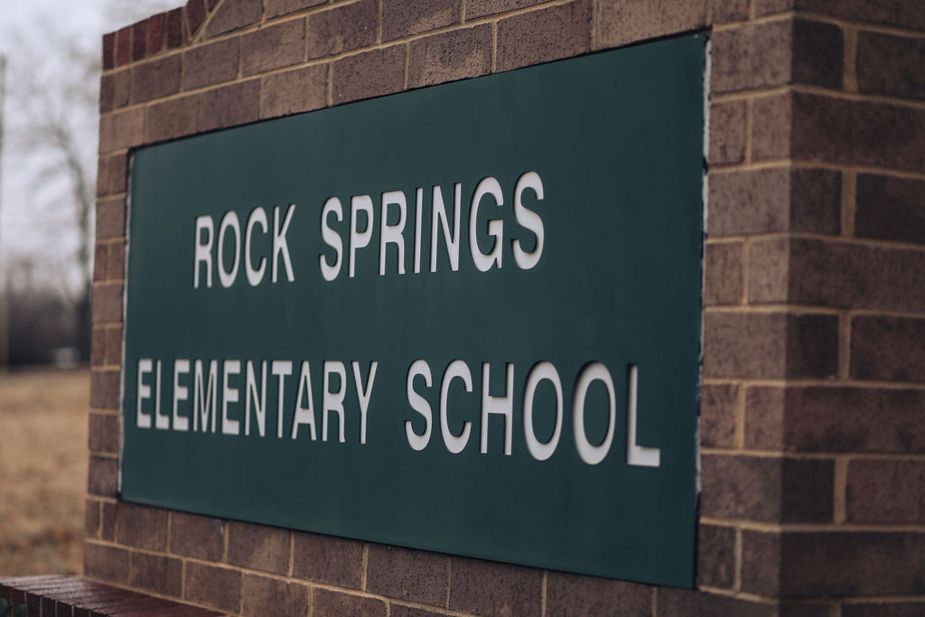 Green sign with white text mounted on a brick wall. Text reads "ROCK SPRINGS ELEMENTARY SCHOOL."