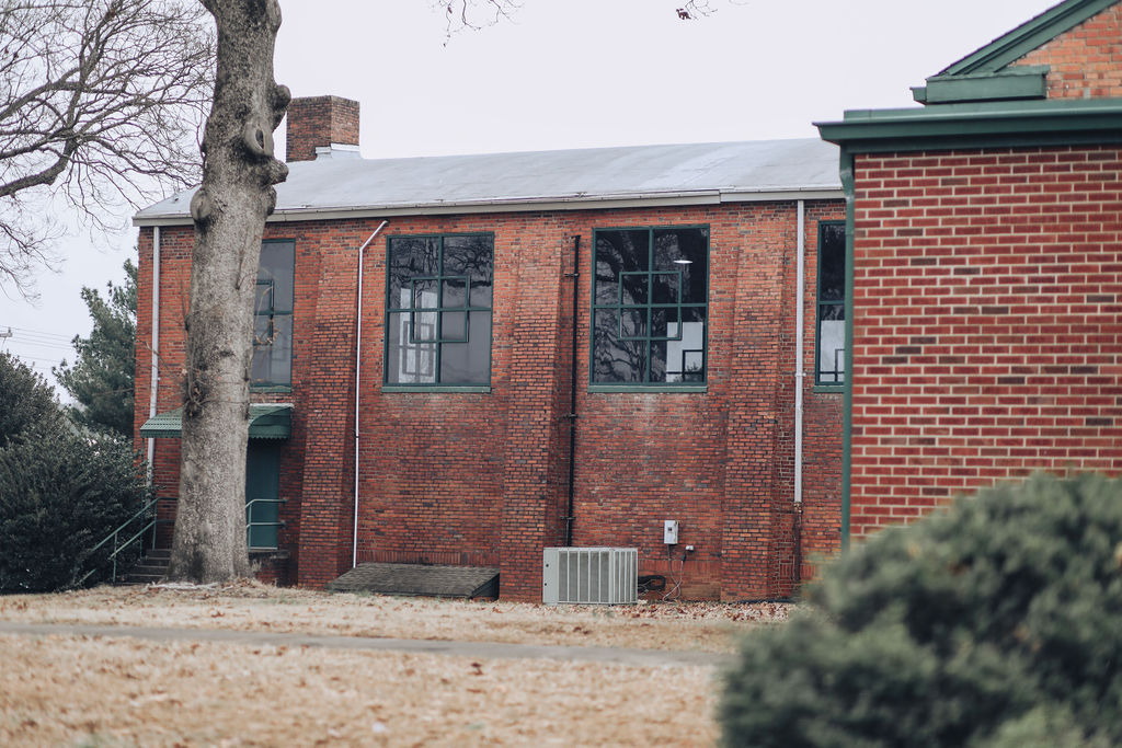 A brick building with a gray roof, three large windows, and an air conditioner unit. Trees and greenery surround the structure.