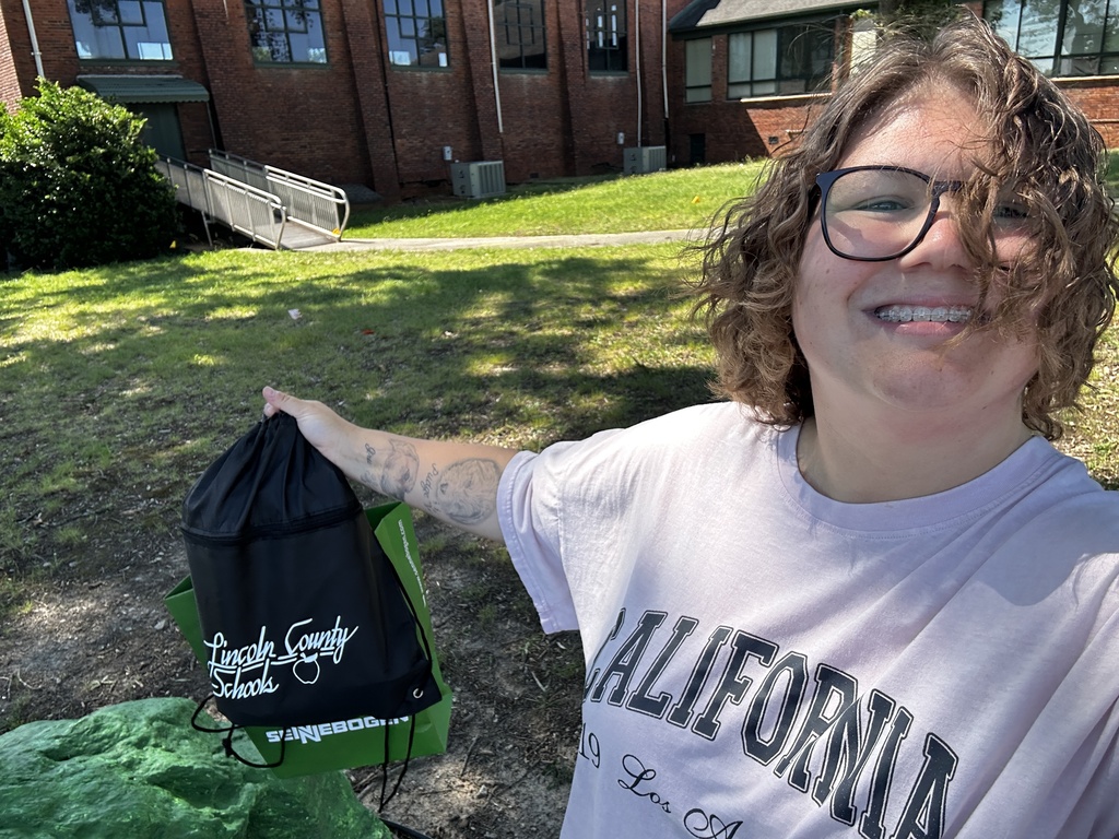 Person smiling and holding a bag in front of a building. Green bin with black bag nearby.