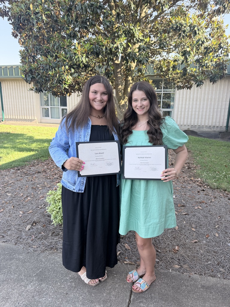 Two women stand on a sidewalk, smiling and holding certificates, in front of a building.